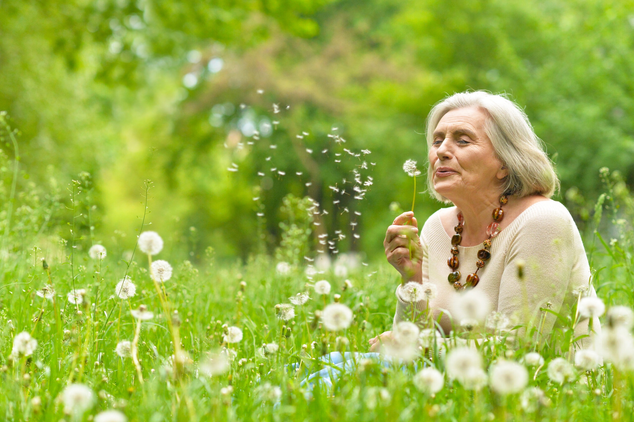 An elderly woman in a field of dandelions gently blows on a dandelion puff, scattering seeds into the air, capturing a moment of joy in nature.